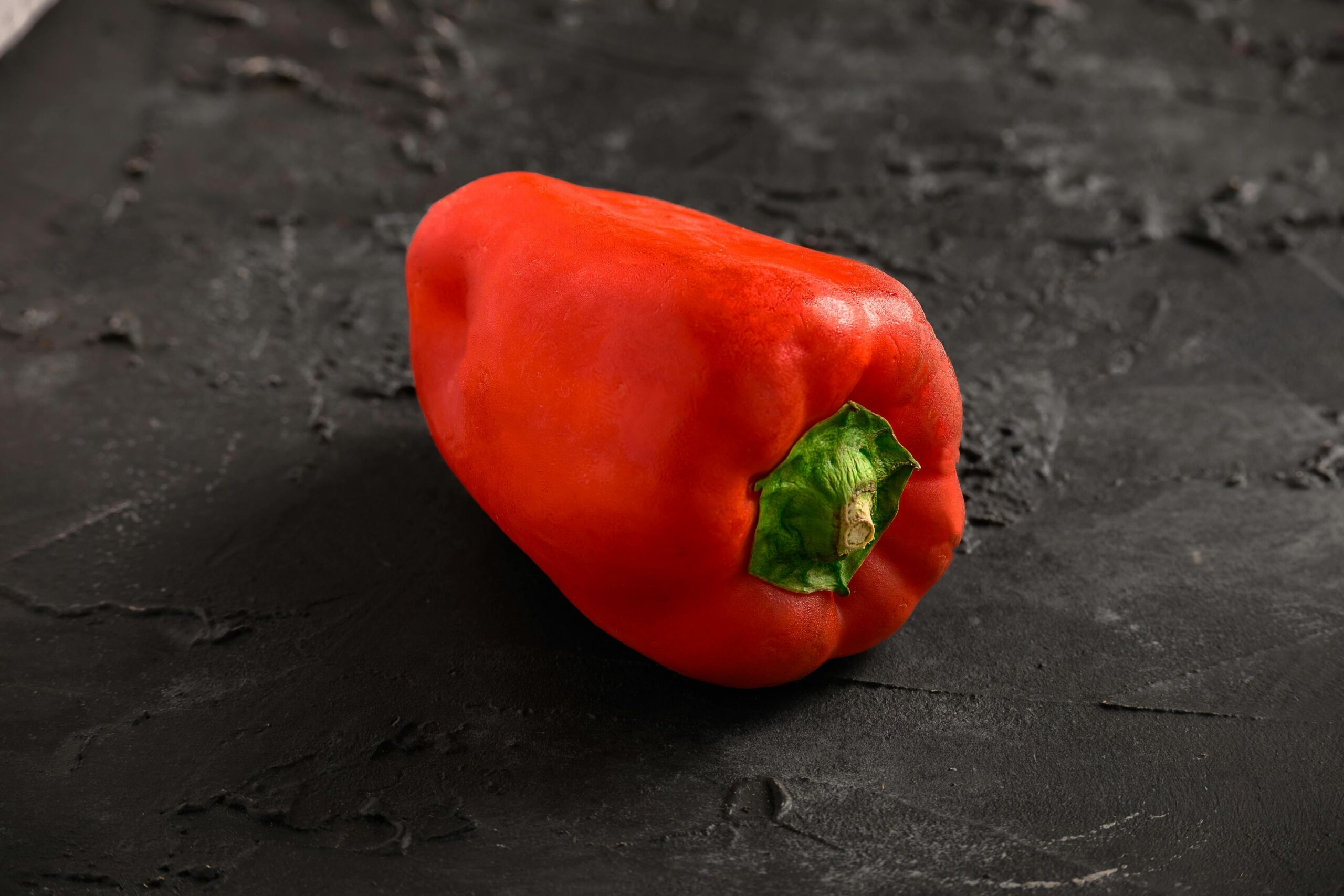 Nahaufnahme einer frischen roten Paprika auf einem strukturierten schwarzen Hintergrund, ideal fÃ¼r die Lebensmittelfotografie.